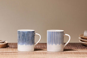Two white nkuku Karuma Ceramic Mug - Large (Set of 2), featuring vertical blue lines, sit side by side on a wooden surface, with handmade mugs, stacked plates, a napkin, and a cork coaster in the neutral beige background.