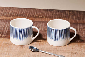 The Karuma Ceramic Mug - Small (Set of 2) by nkuku features handmade Vietnamese-style ceramics with vertical blue lines on the upper half, shown on a rustic wooden surface with a metal spoon and a plain beige background.