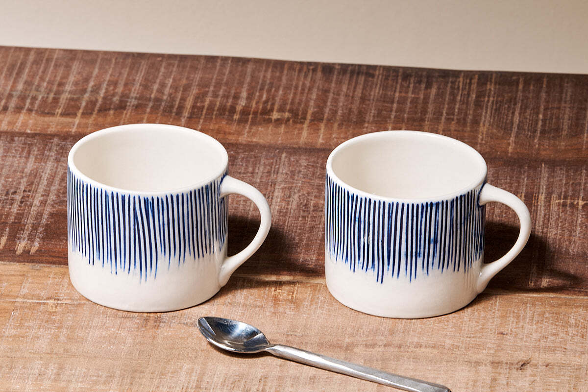 The Karuma Ceramic Mug - Small (Set of 2) by nkuku features handmade Vietnamese-style ceramics with vertical blue lines on the upper half, shown on a rustic wooden surface with a metal spoon and a plain beige background.