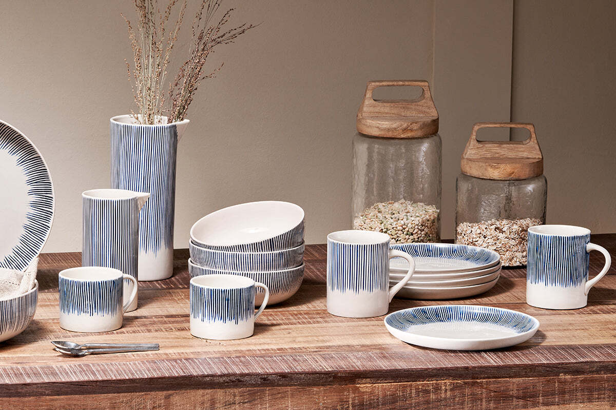 The Karuma Ceramic Mug - Small (Set of 2) by nkuku features white Vietnamese ceramics with blue vertical stripes, handmade design, and pairs well with plates, bowls, and a matching pitcher. Two glass jars with wooden lids sit in the background.