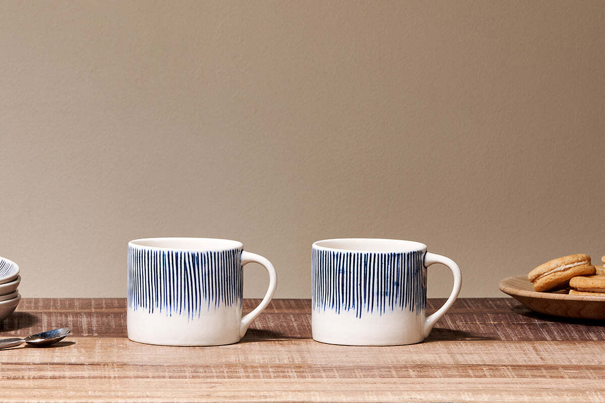A set of two Karuma Ceramic Mugs - Small by nkuku, featuring blue vertical stripes, are placed on a wooden table with a spoon to the left and a small bowl of cookies to the right.