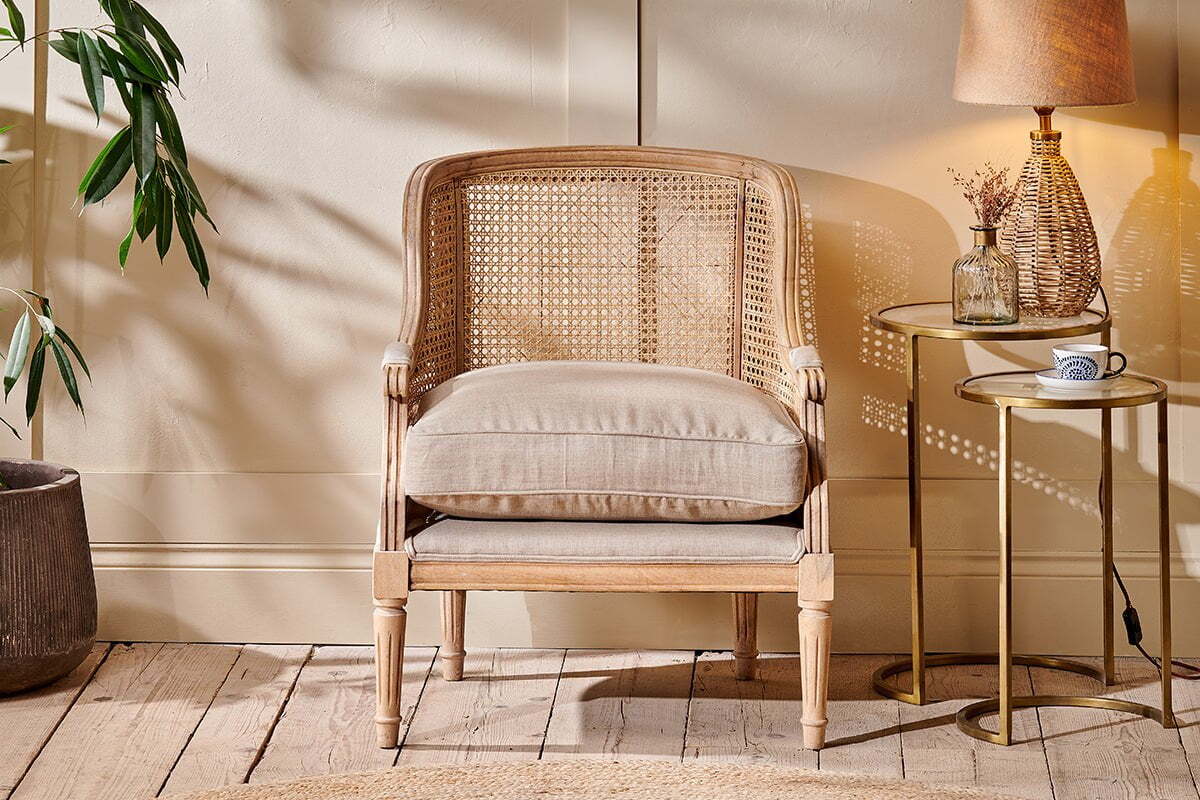 A cozy room features the Kaziria Cane & Linen Armchair - Natural by nkuku with a beige cushion. Nearby, a round side table holds a lamp, glass bottle, and coffee cup. A potted plant sits left as sunlight casts shadows on the cream wall and wood floor.