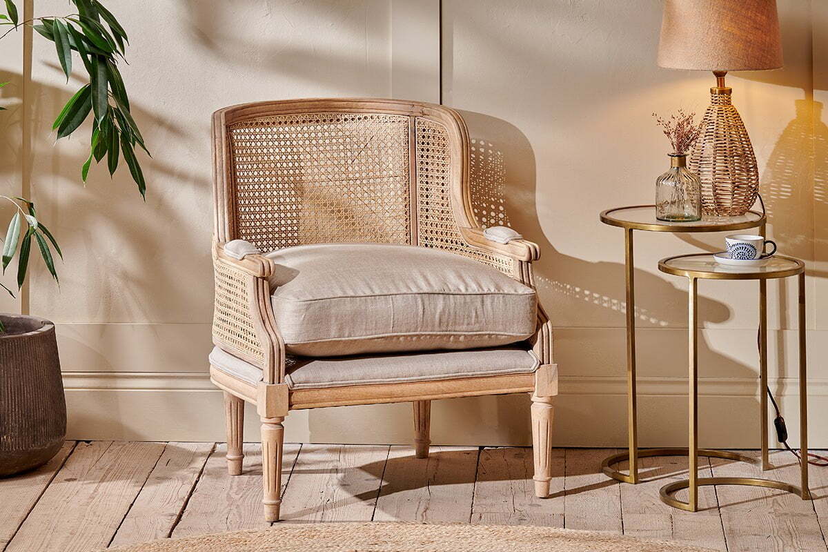 The Kaziria Cane & Linen Armchair by nkuku, featuring a woven back, sides, beige cushion, and armrest pads, sits on a wooden floor beside a round gold side table with a lamp, dried flowers in a vase, and a cup. Sunlight brightens the room and nearby potted plant.