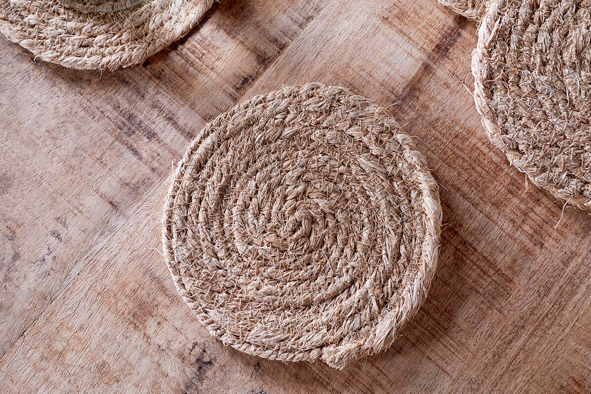 The Keso Woven Coasters - Natural (Set of 4) by nkuku feature a spiral pattern and natural jute fibers. Shown on a light wooden surface, they’re perfect for minimalist table decor, with two more coasters partly visible at the corners.
