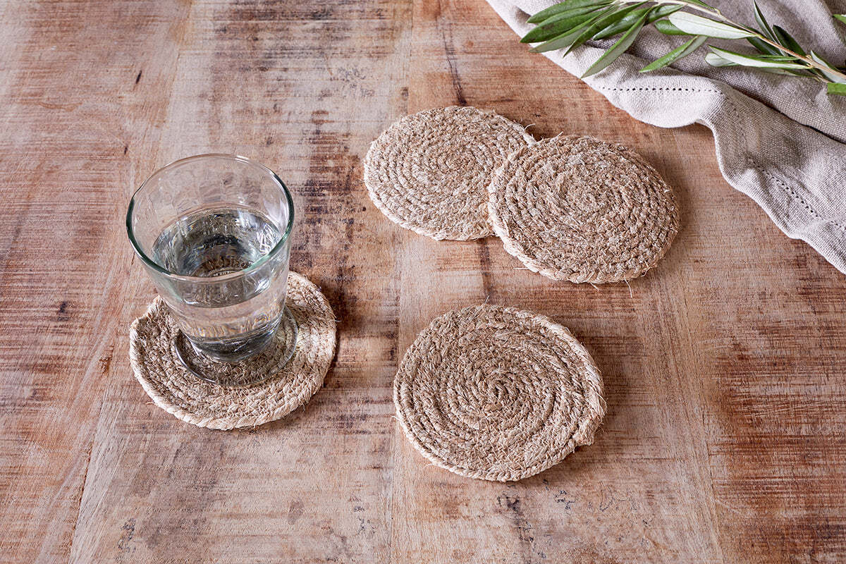 A glass of water sits on an nkuku Keso Woven Coaster—Natural (Set of 4)—atop a rustic wooden table. Three empty coasters are nearby, with a folded beige cloth and greenery sprigs in the upper right corner.