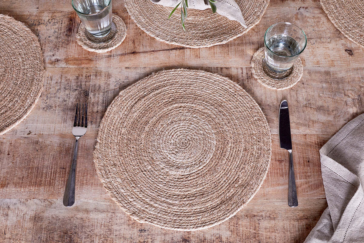 A rustic table features nkuku’s Keso Woven Placemats in natural, paired with a fork, knife, glass of water on a matching coaster, and a beige cloth napkin—all styled for minimalist table decor atop a wooden surface.