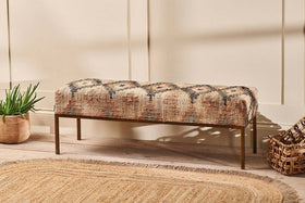 The nkuku Kesu Kilim & Iron Bench, featuring geometric beige, brown, and blue fabric, sits against a cream wall beside a potted plant and woven basket. A round jute rug on the light wood floor completes this warm, inviting scene.