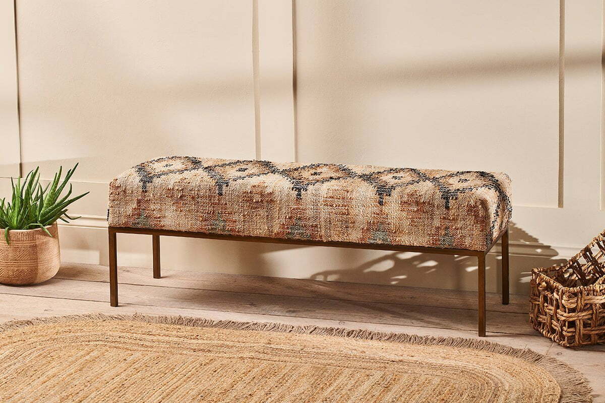 The nkuku Kesu Kilim & Iron Bench, featuring geometric beige, brown, and blue fabric, sits against a cream wall beside a potted plant and woven basket. A round jute rug on the light wood floor completes this warm, inviting scene.