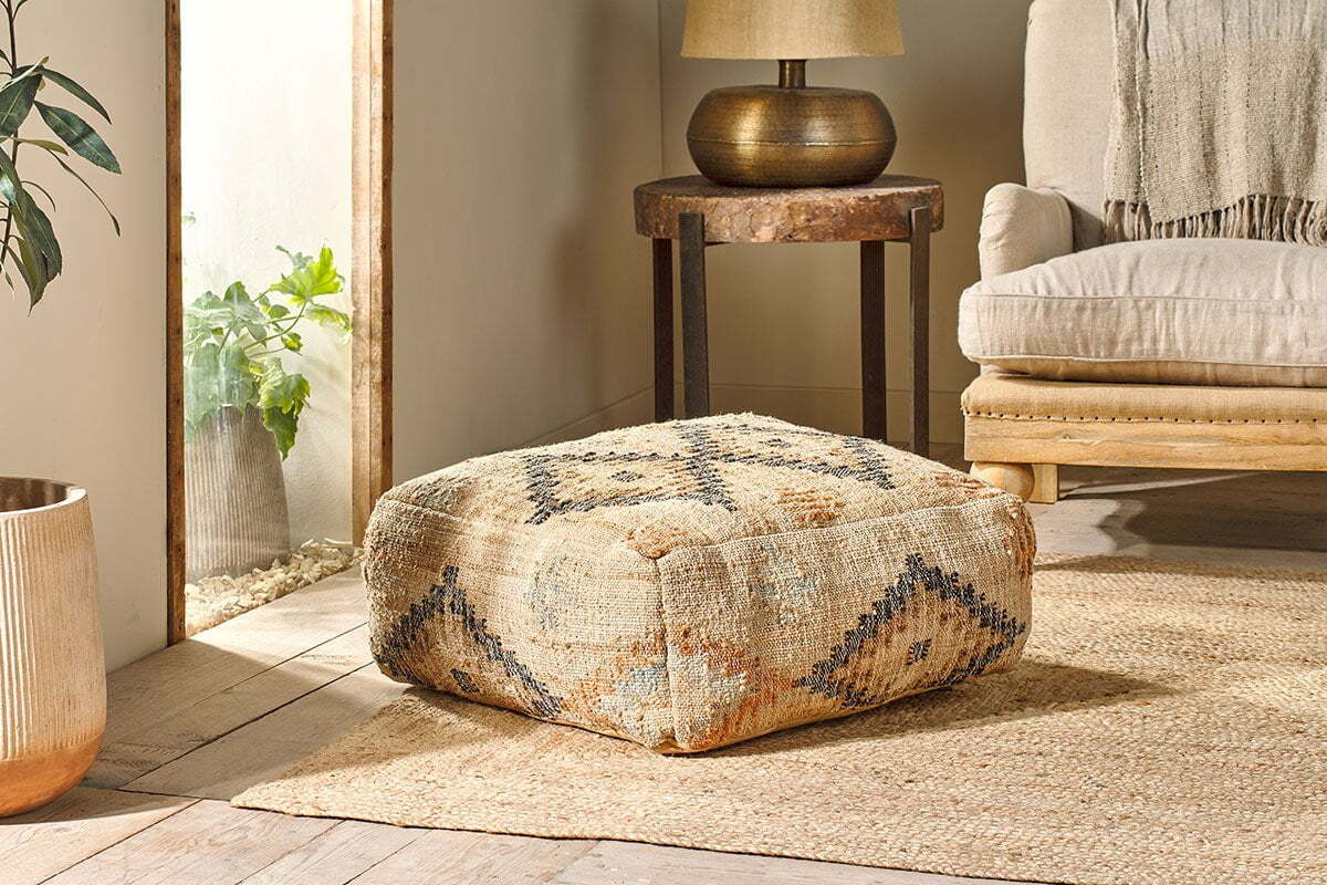 The nkuku Kesu Kilim Pouf - Large, featuring a geometric textured pattern, rests on a woven rug in a sunlit room. Nearby are a chair and a lamp on a side table.