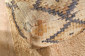 The nkuku Kesu Kilim Pouf - Large, featuring tan, black, and mustard geometric patterns, sits on a natural braided jute rug over light wood floors, accentuating a rustic bohemian vibe with its woven texture and bold design.