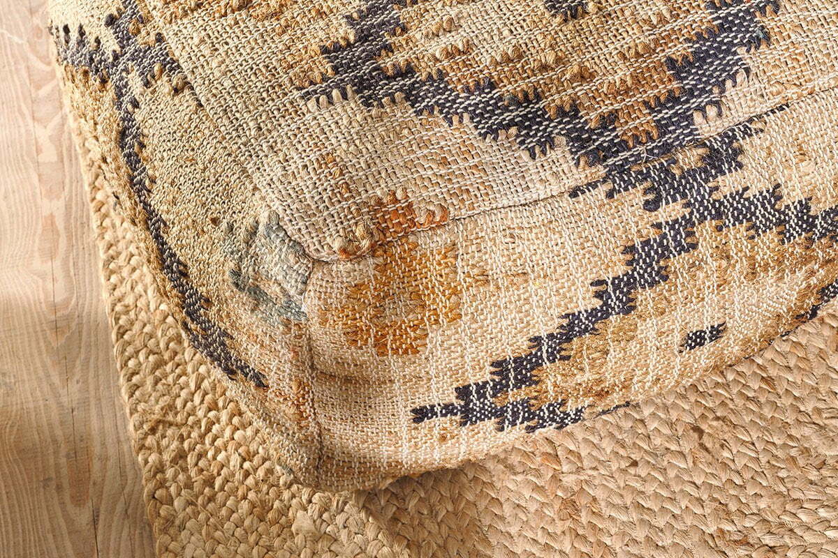 The nkuku Kesu Kilim Pouf - Large, featuring tan, black, and mustard geometric patterns, sits on a natural braided jute rug over light wood floors, accentuating a rustic bohemian vibe with its woven texture and bold design.
