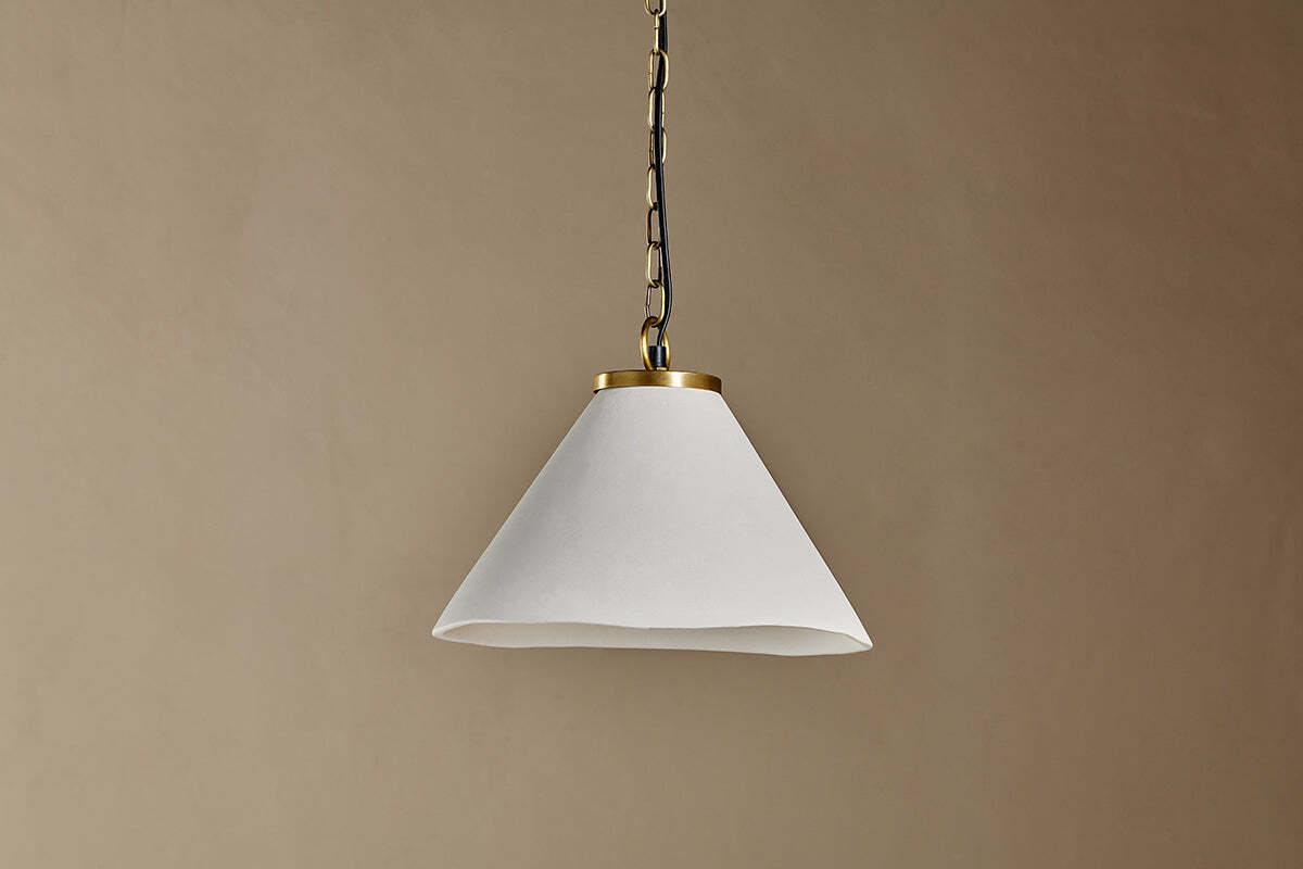 The Kiara Organic Shape Ceramic Pendant Light by nkuku hangs from the ceiling by a chain, featuring an off-white conical shade with smooth finish and antique brass trim—ideal for kitchen lighting against a neutral beige wall.