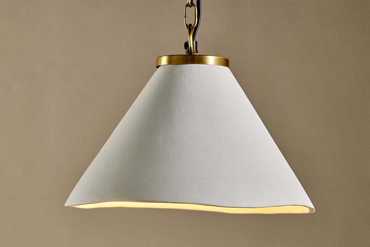 The Kiara Organic Shape Ceramic Pendant Light by nkuku features a wide, wavy-edged off-white shade and antique brass chain, creating a handcrafted modern look against a taupe backdrop.