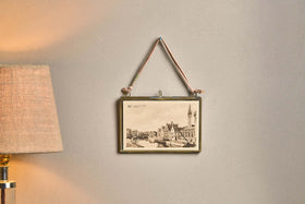 A Kiko Brass Frame by nkuku, displaying a vintage European cityscape postcard, hangs on a light beige wall by a recycled sari tie. Next to it, a textured lampshade softly illuminates the space with a warm glow.
