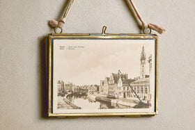 A vintage black-and-white canal scene is displayed in the Kiko Brass Frame by nkuku, which hangs on a beige wall with a pink recycled sari tie. The upper left of the photo reads: “Gent. Quai aux Herbes. Gand.—Graslei.”.