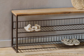 The nkuku Kiyoma Iron & Wood Low Standing Shelves - Natural features a mango wood seat, metal frame, and two wire shelves holding two pairs of sandals for an industrial look against a light wall and wooden floor.
