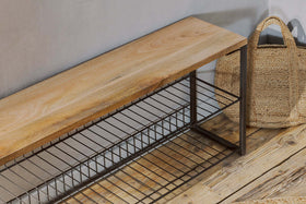 The Kiyoma Iron & Wood Low Standing Shelves by nkuku, featuring mango wood and a metal frame with two wire shelves, stands on a rustic wooden floor beside a straw tote bag, bringing an industrial touch to the space.