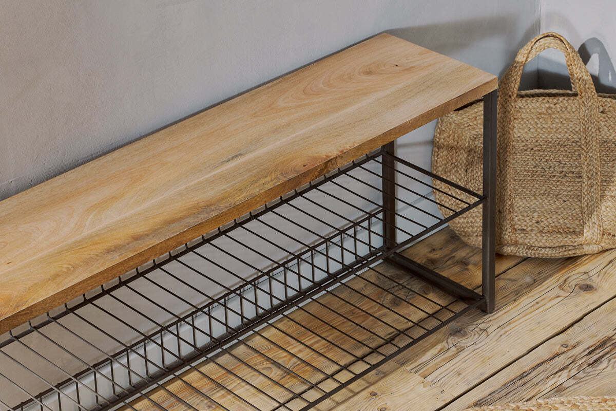 The Kiyoma Iron & Wood Low Standing Shelves by nkuku, featuring mango wood and a metal frame with two wire shelves, stands on a rustic wooden floor beside a straw tote bag, bringing an industrial touch to the space.