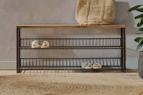 The nkuku Kiyoma Iron & Wood Low Standing Shelves - Natural features a mango wood seat, metal frame, and two wire shelves holding shoes. A woven bag rests on top beside a round rug, with a potted plant by the light-colored wall.