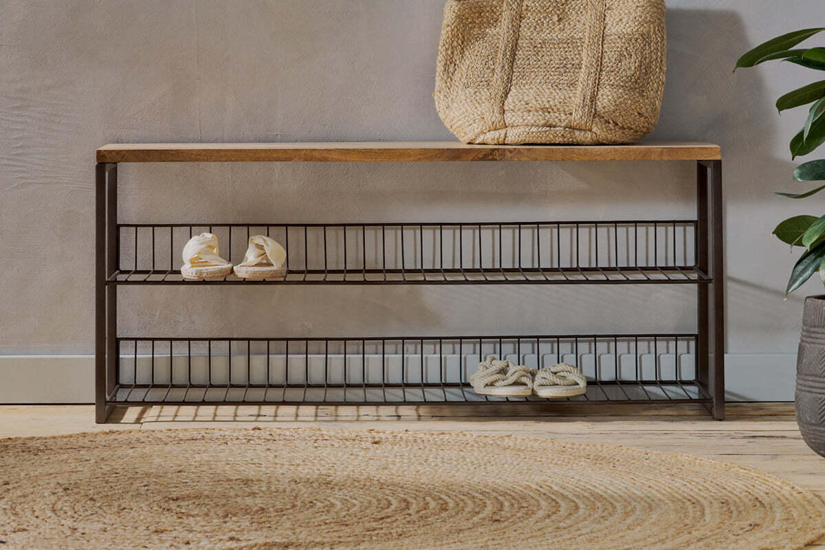 The nkuku Kiyoma Iron & Wood Low Standing Shelves - Natural features a mango wood seat, metal frame, and two wire shelves holding shoes. A woven bag rests on top beside a round rug, with a potted plant by the light-colored wall.
