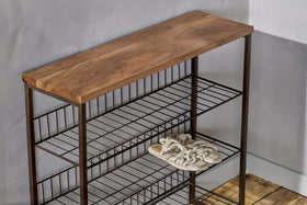 The Kiyoma Iron & Wood Tall Standing Shelves by nkuku, featuring a natural mango wood top and metal frame, stands against a light gray wall. Only a single pair of rope-strap sandals sits on the middle shelf; the others are empty on the wooden floor.