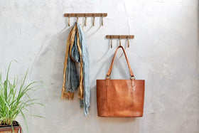 Two Kuna Iron Hooks in Antique Brass by nkuku adorn a textured gray wall. The left hook displays blue and gold scarves, while the right holds a tan leather tote—perfect for organizing small items in style.