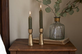 Two green candles in nkukus Lokoja Candle Holder - Antique Brass (Set of 2) sit on wood beside a ribbed glass vase with eucalyptus and a closed book, creating a cozy, rustic decor scene.