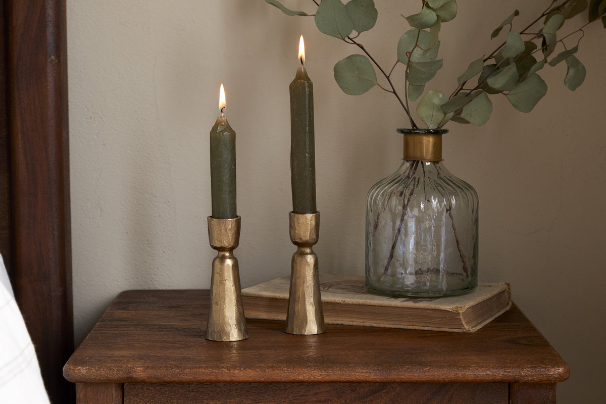Two green candles in nkukus Lokoja Candle Holder - Antique Brass (Set of 2) sit on wood beside a ribbed glass vase with eucalyptus and a closed book, creating a cozy, rustic decor scene.