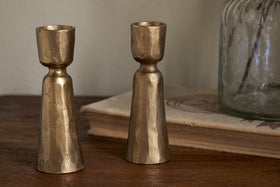 The Lokoja Candle Holder - Antique Brass (Set of 2) by nkuku features gold-toned textured metal, adding an elegant touch to your table when placed beside a book and a glass jar.