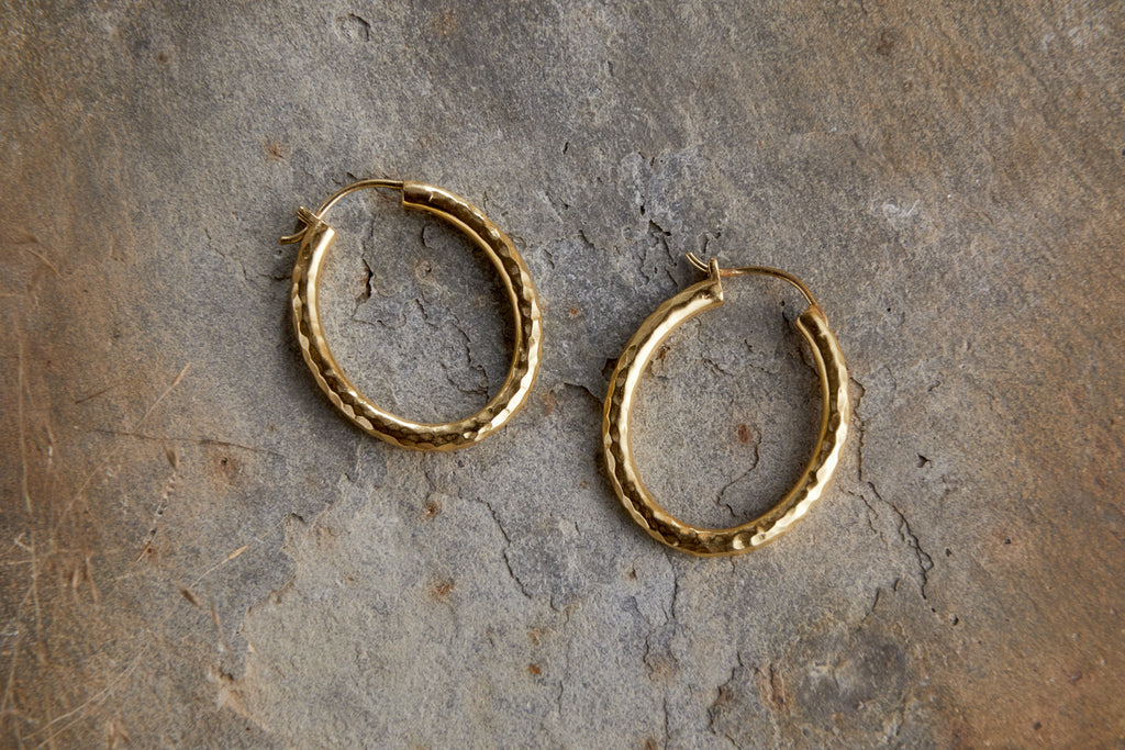 The Labani Earrings - Gold by nkuku, featuring a textured hoop design, are displayed on a gray stone surface, highlighting their timeless elegance.
