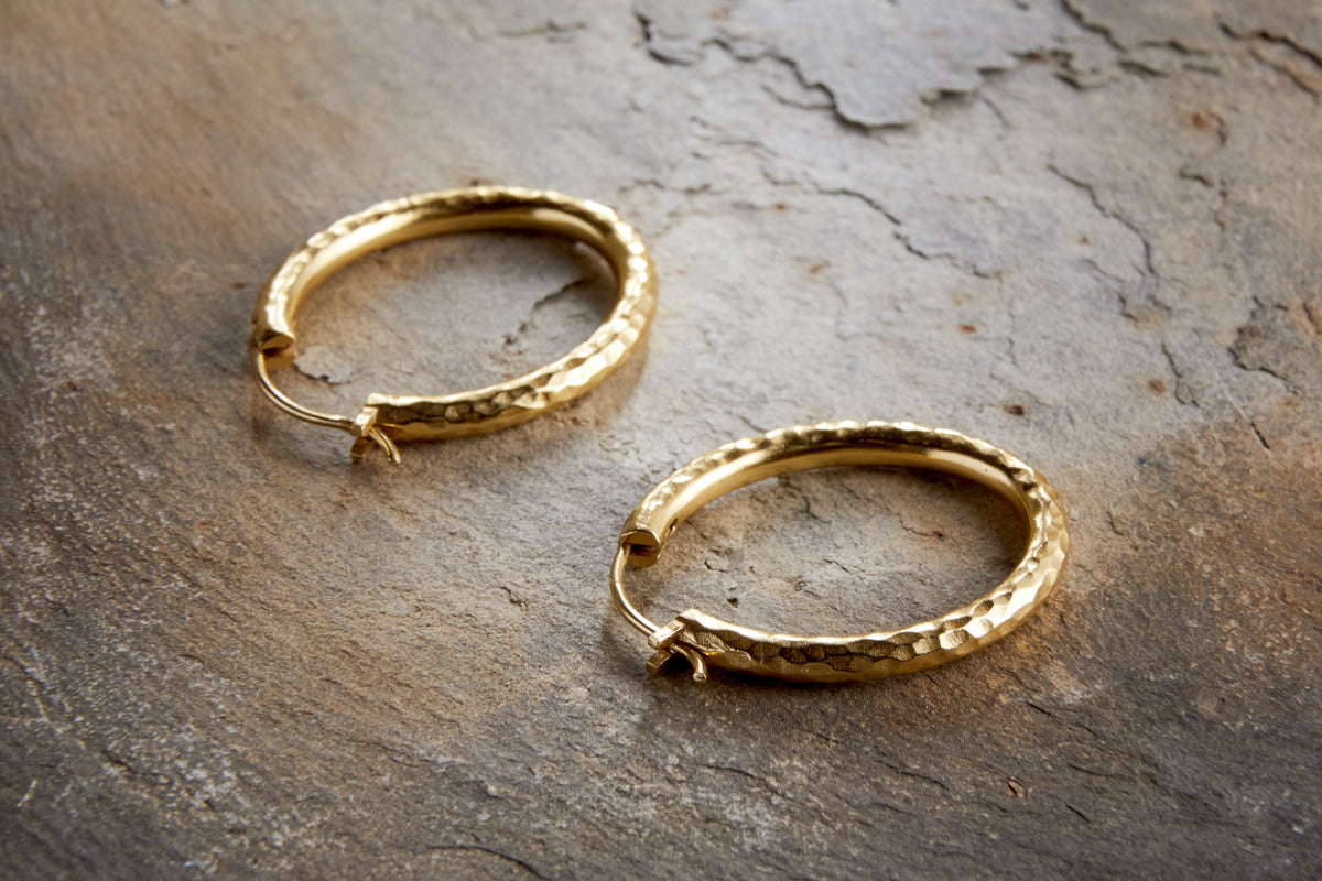 The Labani Earrings - Gold by nkuku, with a textured, hammered finish and slightly open clasps, rest on a rugged stone-like surface—timeless jewellery highlighted by the light.