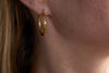 A close-up of a persons ear wearing nkukus Labani Earrings - Gold. Light brown hair frames the ear, and the skin shows a natural complexion with faint freckles.