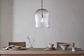 A minimalist dining table with wooden chairs is set with ceramic dinnerware and a linen napkin. On the table stands a small vase of dried flowers. Above, nkuku’s Lumina Fluted Recycled Glass Pendant in clear hangs against a textured white wall.
