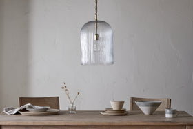 A minimalist dining table with two wooden chairs is set with plates, bowls, a napkin, and a vase of dried flowers. Overhead hangs the nkuku Lumina Fluted Recycled Glass Pendant in clear against a plain textured wall.