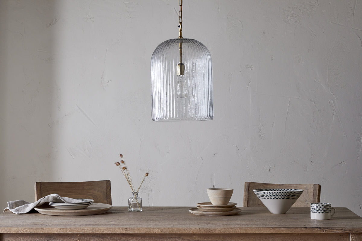 A minimalist dining table with two wooden chairs is set with plates, bowls, a napkin, and a vase of dried flowers. Overhead hangs the nkuku Lumina Fluted Recycled Glass Pendant in clear against a plain textured wall.
