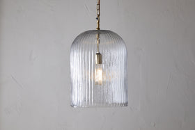 The nkuku Lumina Fluted Recycled Glass Pendant - Clear features a ribbed, dome-shaped shade and brass fixture with a rust-colored cable, casting a gentle glow against a light textured wall.