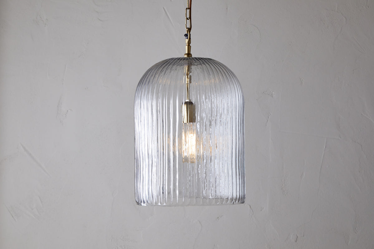The nkuku Lumina Fluted Recycled Glass Pendant - Clear features a ribbed, dome-shaped shade and brass fixture with a rust-colored cable, casting a gentle glow against a light textured wall.