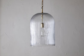 The nkuku Lumina Fluted Recycled Glass Pendant in clear features a ribbed, transparent shade and a rust-coloured fabric cable hanging from a brass fixture, showcasing the bulb against a textured white wall.