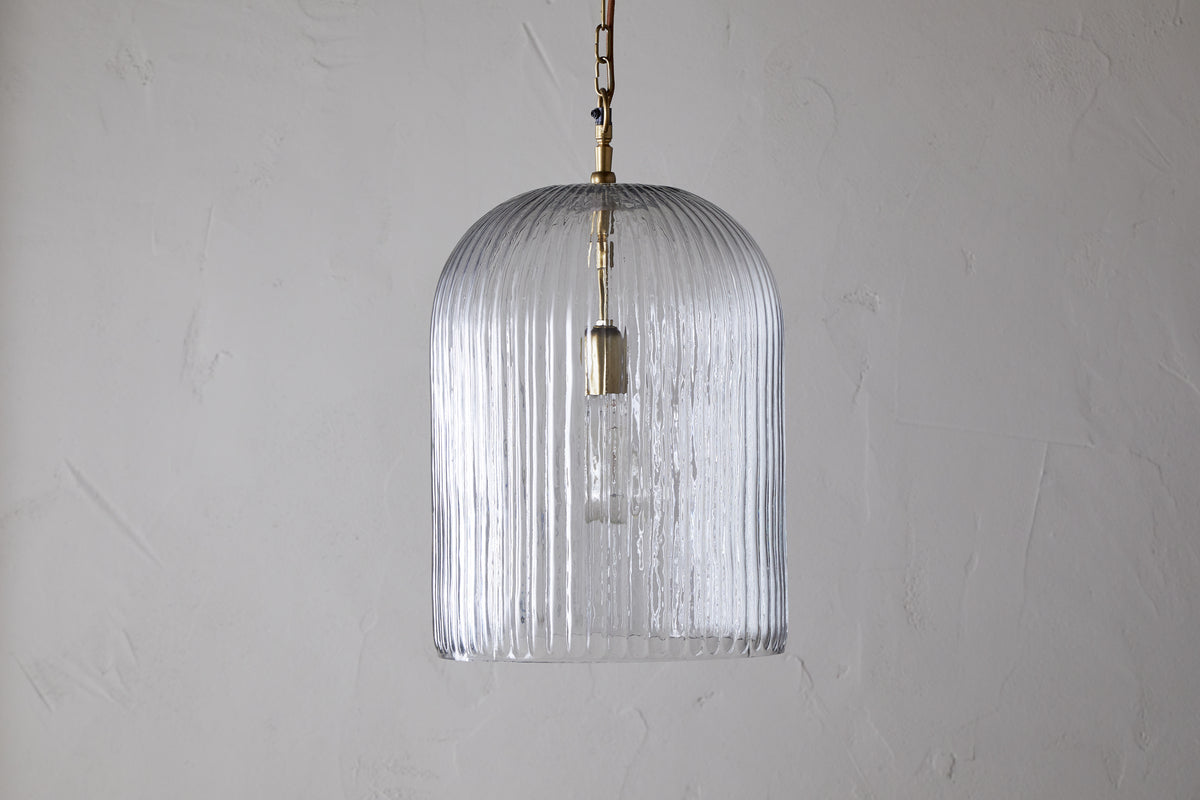 The nkuku Lumina Fluted Recycled Glass Pendant in clear features a ribbed, transparent shade and a rust-coloured fabric cable hanging from a brass fixture, showcasing the bulb against a textured white wall.