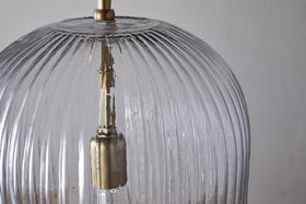 Close-up of the nkuku Lumina Fluted Recycled Glass Pendant in clear, featuring a brass fixture and rust-coloured fabric cable. Vertical grooves accent the transparent shade, with a neutral backdrop highlighting its textured design.