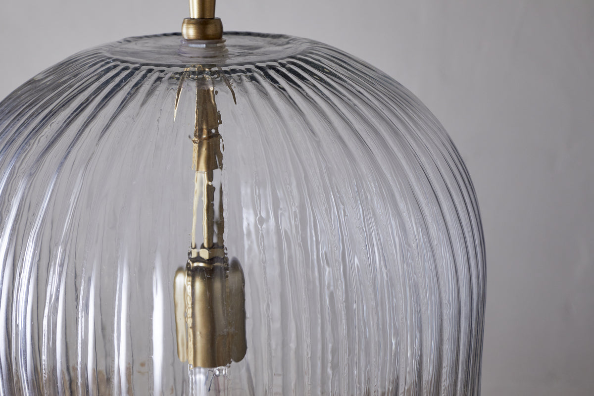 Close-up of the nkuku Lumina Fluted Recycled Glass Pendant in clear, featuring a brass fixture and rust-coloured fabric cable. Vertical grooves accent the transparent shade, with a neutral backdrop highlighting its textured design.