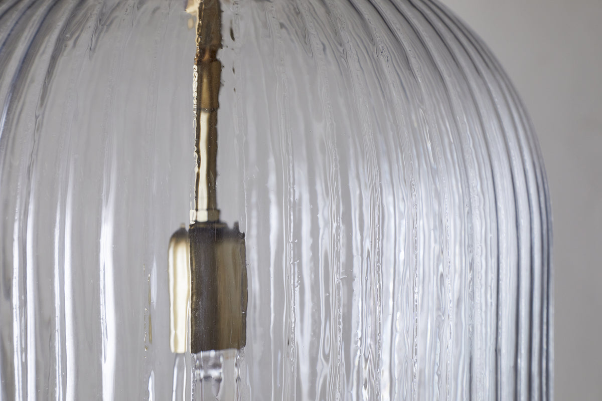 Close-up of the Lumina Fluted Recycled Glass Pendant - Clear by nkuku. The vertical lines and metallic fixture inside stand out against a neutral background, highlighting the pendant's clear recycled glass texture and detail.
