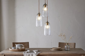A rustic wooden table set with ceramic mugs and plates sits beneath the nkuku Lumina Fluted Recycled Glass Cluster Light—Clear, casting an ambient glow on a textured off-white wall. A vase of dried flowers and a folded napkin complete the scene.