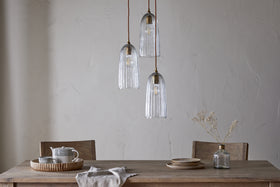A wooden dining table for two is set with plates, a vase of dried white flowers, and a tray with a teapot and mugs. Overhead, the nkuku Lumina Fluted Recycled Glass Cluster Light - Clear casts an ambient glow on the light-colored wall.