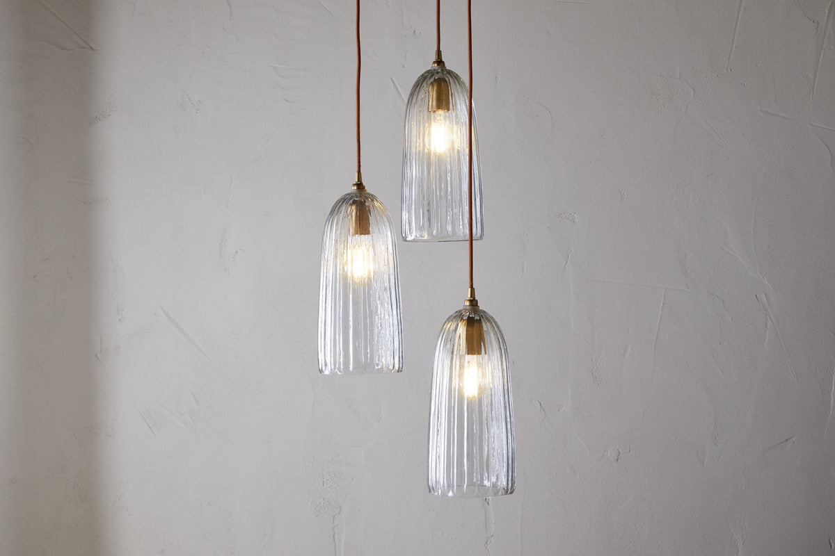 The nkuku Lumina Fluted Recycled Glass Cluster Light features three clear, ribbed glass pendants hung at different heights from brown cords, casting an ambient glow that enhances the elegant and minimalist design.