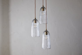 The nkuku Lumina Fluted Recycled Glass Cluster Light - Clear features three fluted glass pendants with visible bulbs and brass accents, suspended at varying heights from brown cords for a warm, inviting glow.