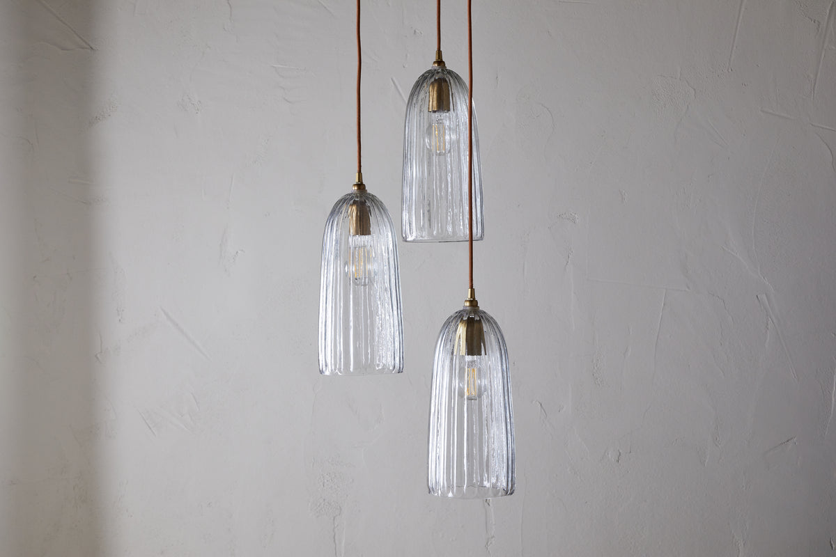 The nkuku Lumina Fluted Recycled Glass Cluster Light - Clear features three fluted glass pendants with visible bulbs and brass accents, suspended at varying heights from brown cords for a warm, inviting glow.