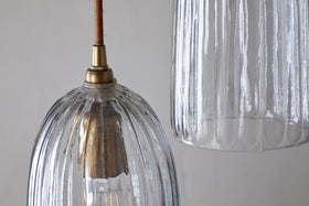Close-up of two Lumina Fluted Recycled Glass Cluster Lights by nkuku, each with clear ribbed glass shades and brass fittings. The left pendant displays a visible bulb glowing, while the right one is partly viewed against a light gray background.