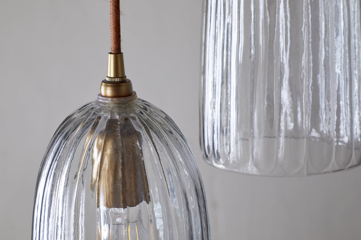 Close-up of two Lumina Fluted Recycled Glass Cluster Lights by nkuku, each with clear ribbed glass shades and brass fittings. The left pendant displays a visible bulb glowing, while the right one is partly viewed against a light gray background.