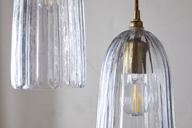 The Lumina Fluted Recycled Glass Cluster Light by nkuku features two clear, ribbed pendants with brass fittings. Visible bulbs cast a warm glow, accentuating the textured glass and metallic accents against a soft, neutral backdrop.